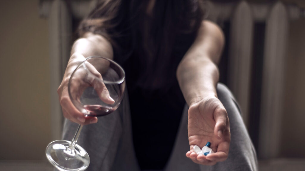 Woman sitting on floor with wine in one hand and benzodiazepines in the other hand.